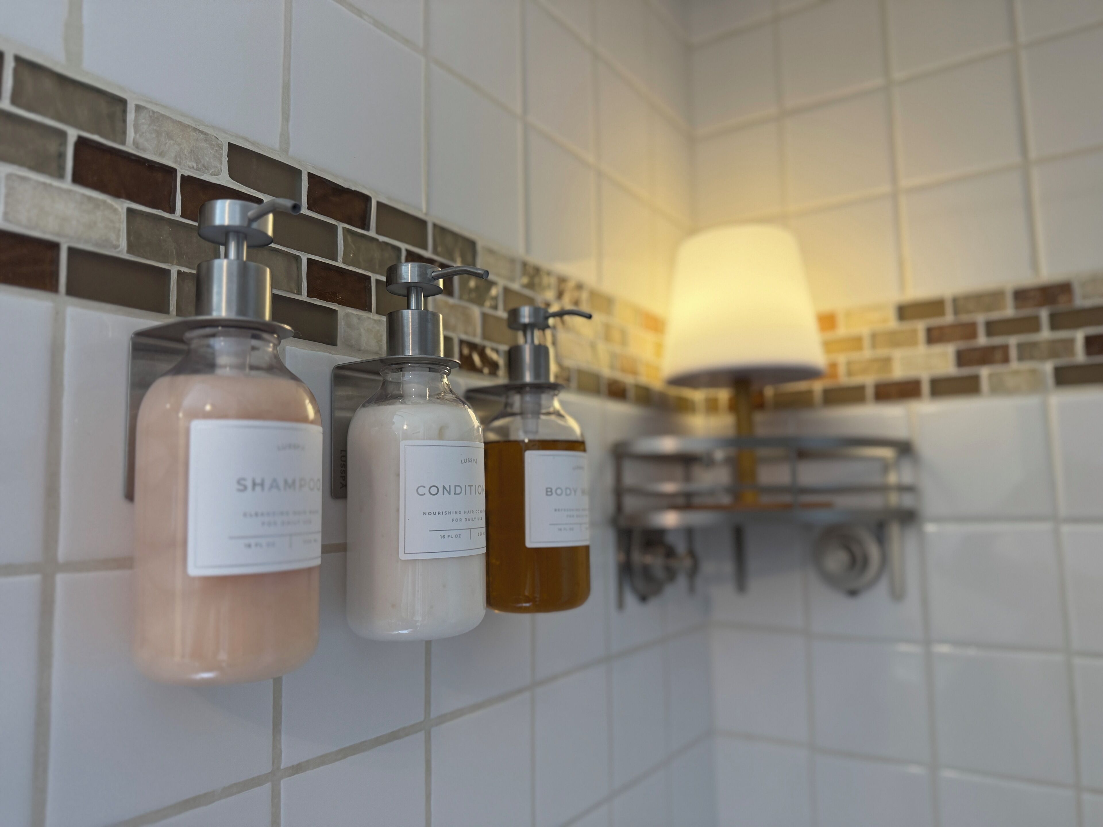 A bathroom with a tiled wall, a hanging lamp, and three bottles of shampoo and conditioner.