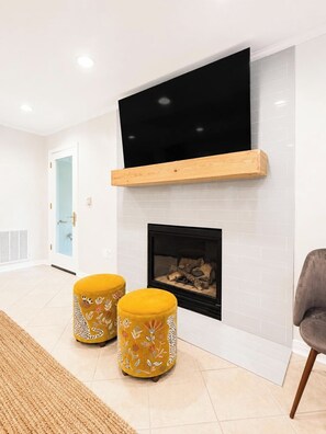 A modern living room with a flat-screen TV mounted above a fireplace, two decorative stools, and a grey armchair. - The Park House, 1.8mi to Square, VelvetDitchVillas (Oxford)