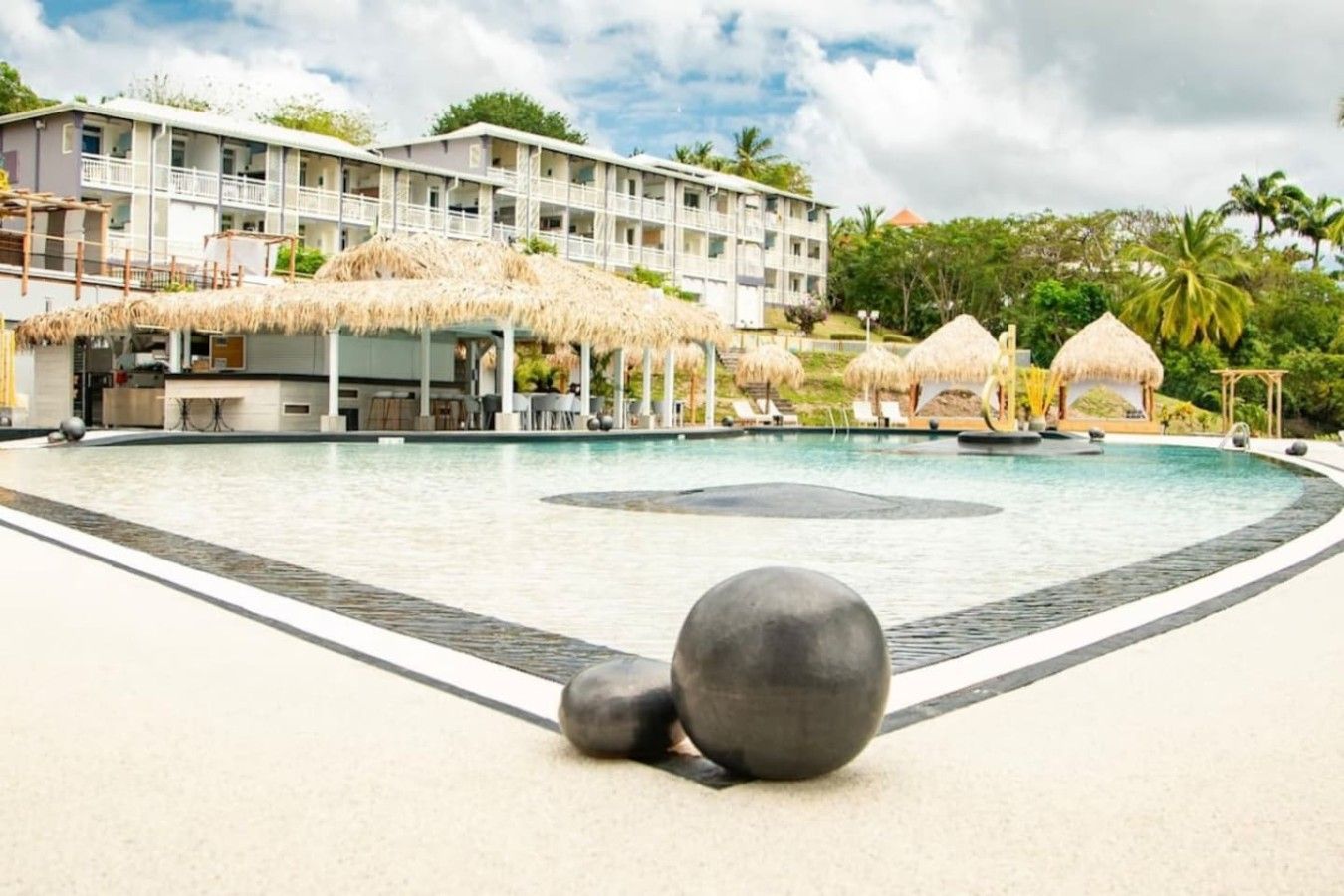 Un área de piscina en un resort, con una piscina grande, sillas de descanso y estructuras de techo de paja.