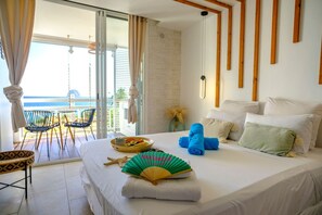 Une chambre d’hôtel avec un lit, un ventilateur, un plateau de fruits et une vue sur la mer depuis le balcon.
