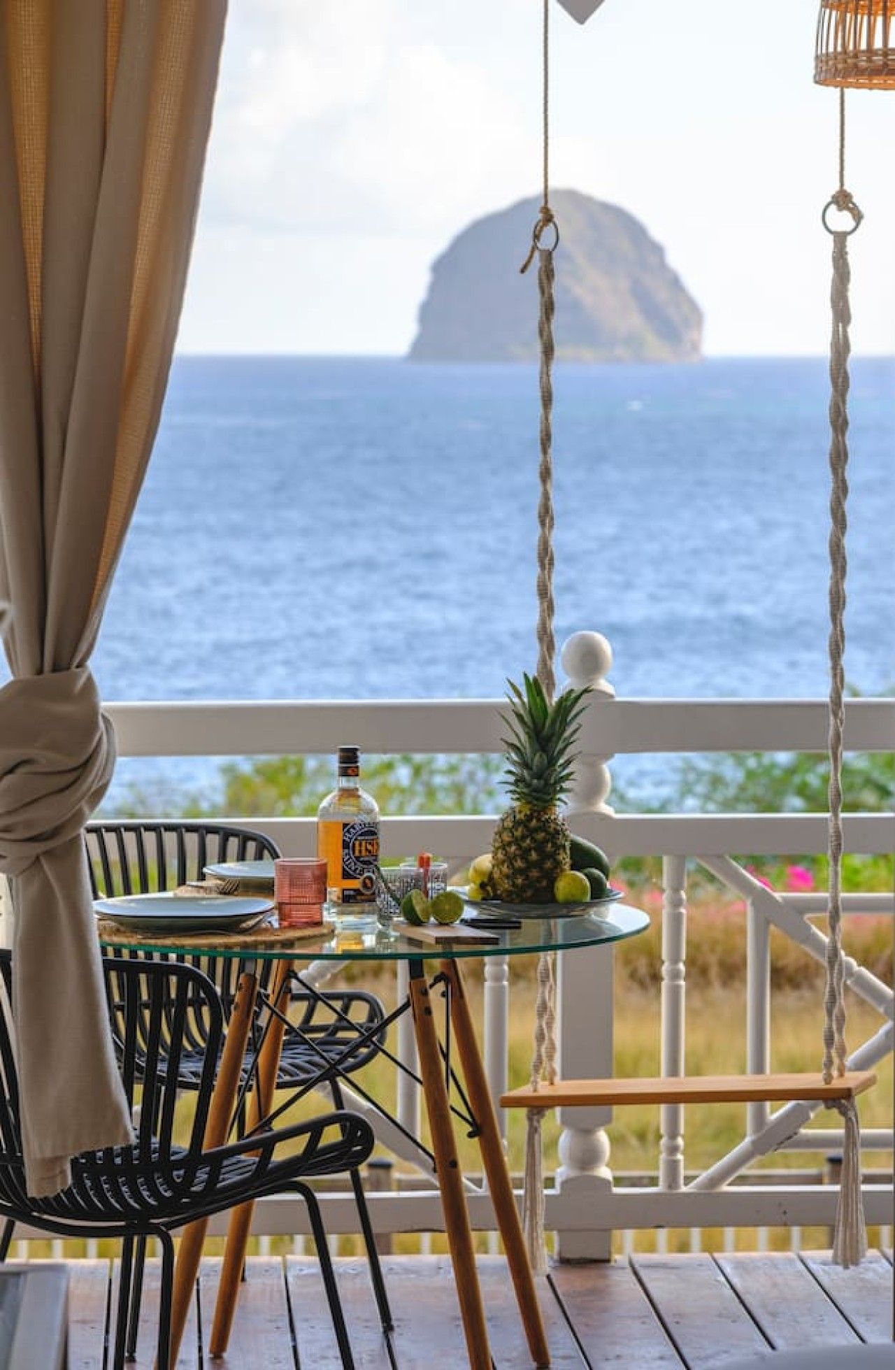 Une table en verre avec une bouteille et des fruits, sur un balcon donnant sur la mer.