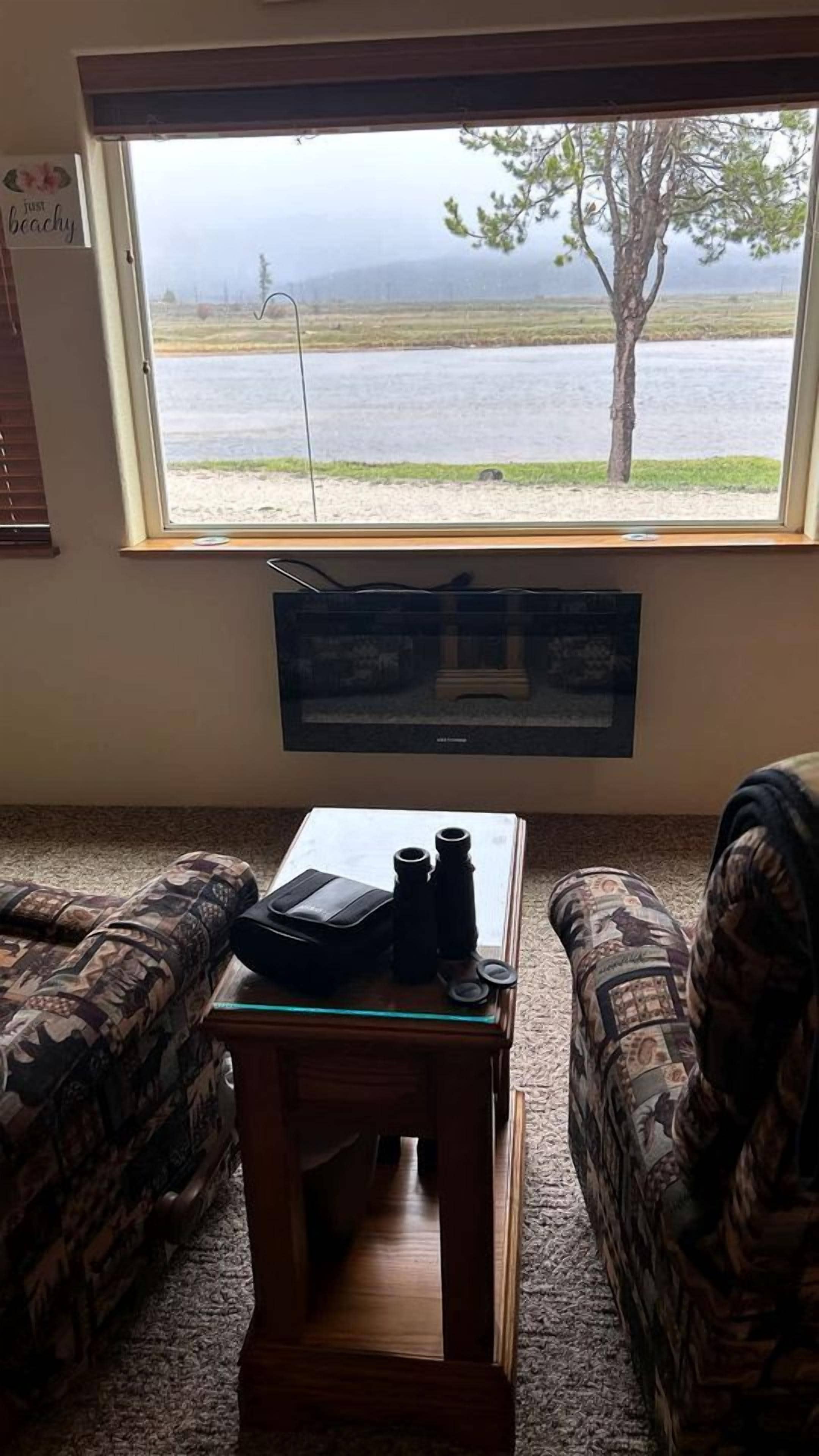 Ein Zimmer mit großem Fenster mit Blick auf ein Gewässer, einem an der Wand befestigten Fernseher und zwei gemusterten Sesseln.