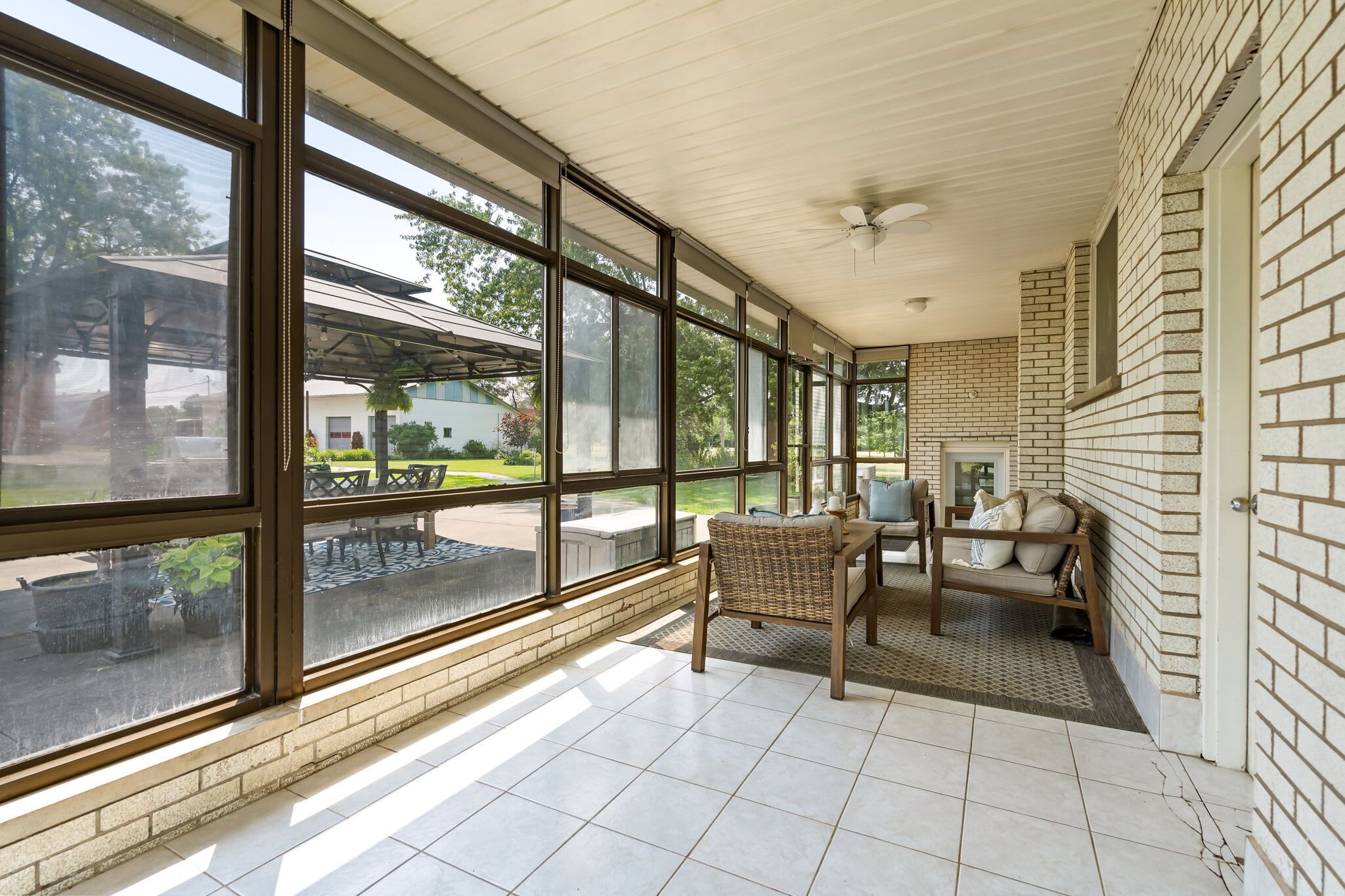 Ein überdachter Patio-Bereich mit großen Fenstern, einem Deckenventilator und einer Sitzecke mit einer Couch und Sesseln.