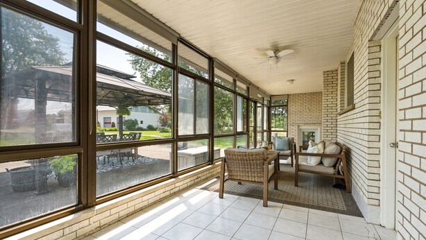 Un patio techado con amplios ventanales, un ventilador de techo y un área de estar con sofá y sillones.