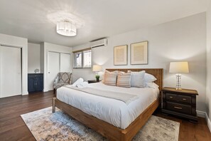 Ein Schlafzimmer mit einem hölzernen Bett, Nachttischen, einem Beistelltisch, einer Deckenleuchte und einem Fenster mit Jalousien.