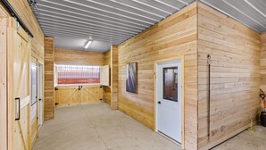 Un espacio interior amplio con paredes de madera, suelo de concreto y techo de metal. Hay puertas y ventanas, una de ellas ofrece vistas al exterior.