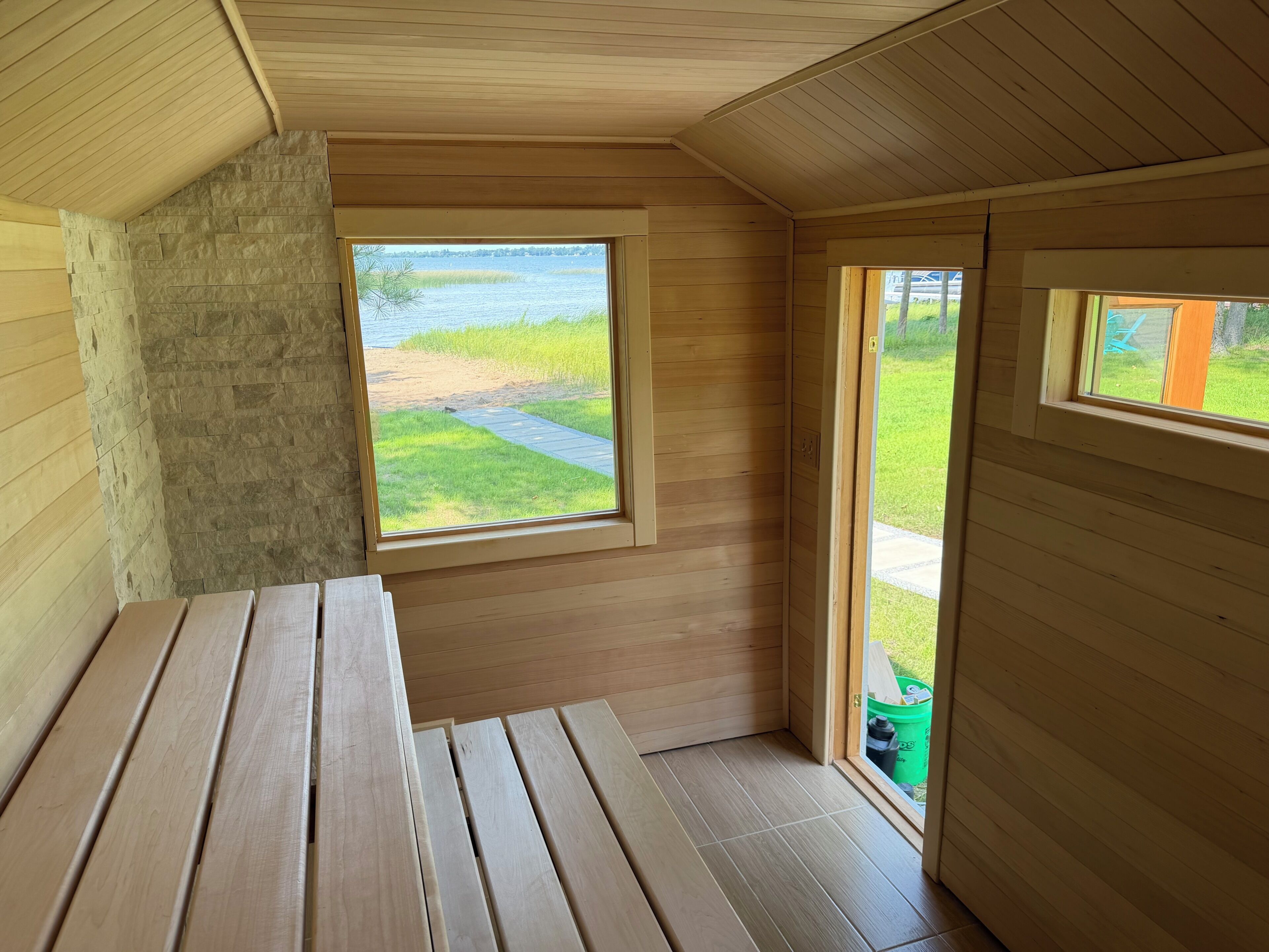 A wooden interior with a bench, a window with a view of a grassy area and water, and a door leading outside.