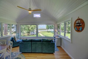 A room with a large window, a ceiling fan, a blue sofa, and a wooden sign on the wall. - Stunning Sunset Views on Round Lake - 3BR Lakefront Cabin with Sauna (Brainerd)