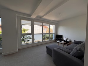 Ein modernes Wohnzimmer mit großem Fenster, einem grauen Sofa, einem kleinen weißen Tisch und einem Flachbildfernseher.