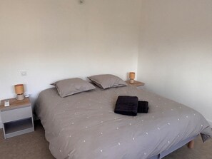 A single bed with a grey duvet and pillows, a bedside table with a lamp, and a folded black towel on the bed. - T4 Neuf & Climatisé - Au coeur de St Paul les Dax (Saint-Paul-lès-Dax)