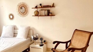 Un dormitori amb un llit, una cadira bressol, una taula petita amb una làmpada i dues prestatgeries murals amb objectes decoratius.