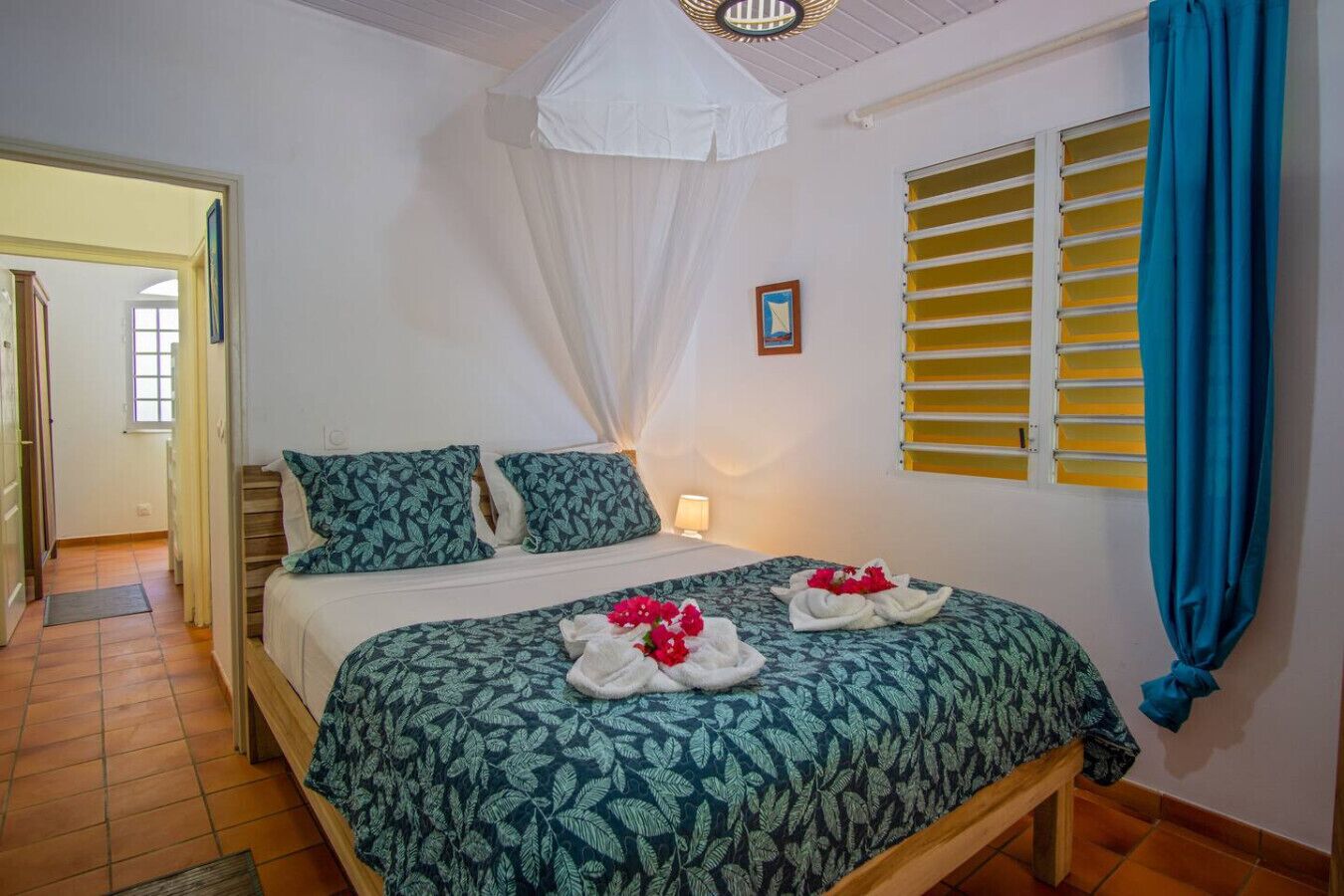 A hotel room with a bed, a bedside table, a window with yellow blinds, and a door leading to another room.