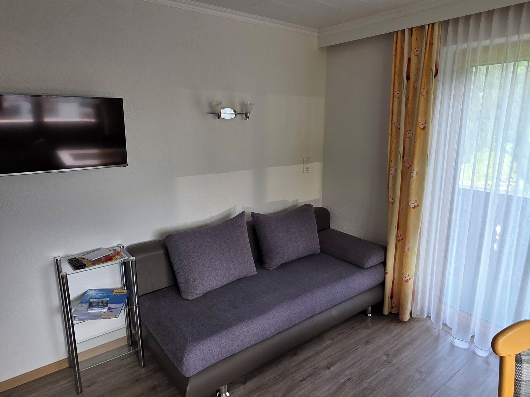 A modern living room with a grey sofa, a flat-screen TV mounted on the wall, and a small side table with books.