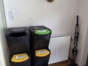 Zwei Müllbehälter, einmal für Papier und einmal für Metall, sowie ein Staubsauger in einem Raum mit einem Heizkörper.