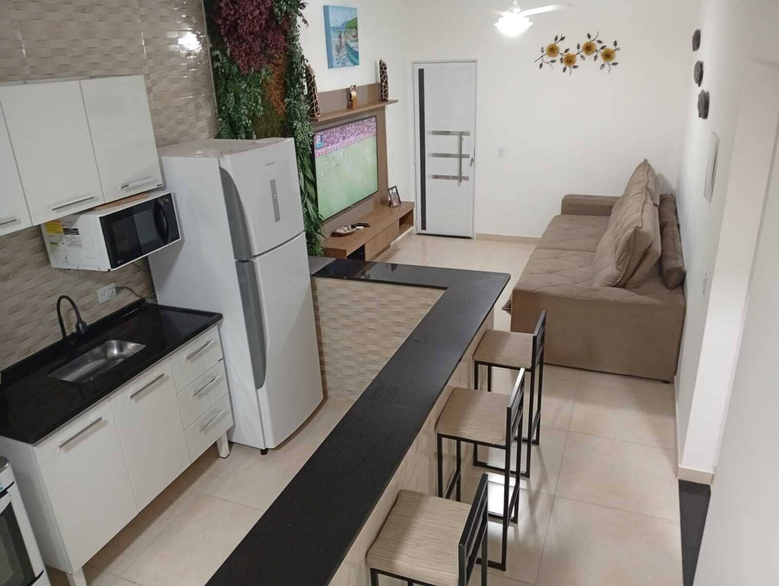 Eine kompakte Küche mit weißen Schränken, einem Kühlschrank, einer Mikrowelle und einem Spülbecken. Es gibt ein beiges Sofa und einen Esstisch mit Stühlen. Eine Zierpflanze ist in der Ecke sichtbar.