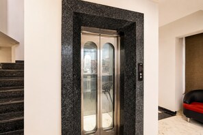 Un ascensor moderno con puerta de vidrio, ubicado entre una escalera y una zona de descanso.