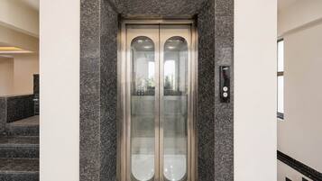 A modern elevator with a metallic finish and glass panels, located in a building with a stone facade and a staircase.