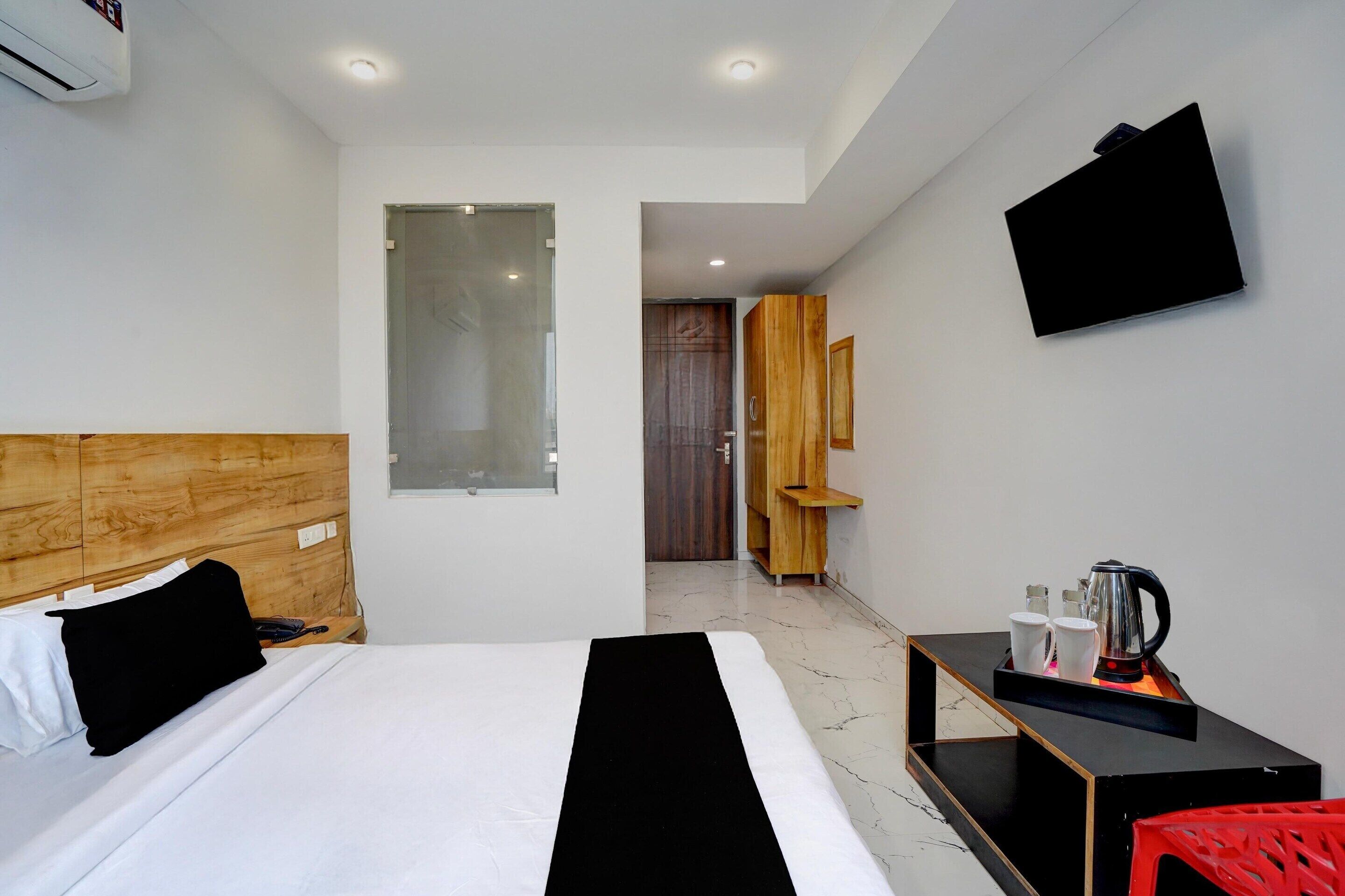A hotel room with a bed, a bedside table with a kettle, a TV mounted on the wall, and a wooden headboard.