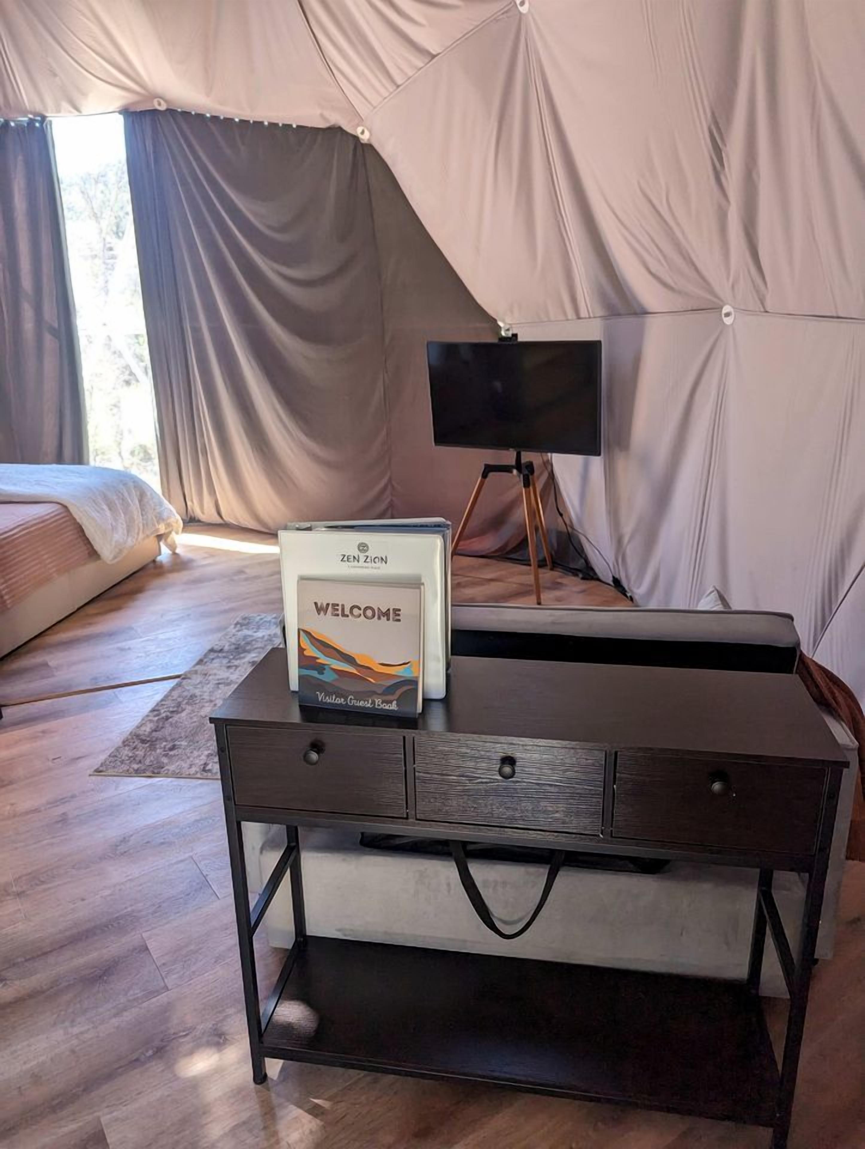 A tented room with a bed, a desk with a welcome sign, and a television on a tripod.