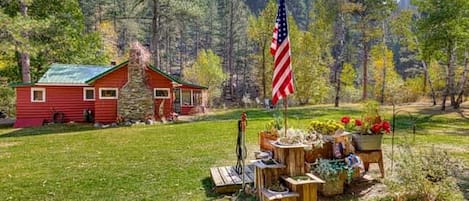 Una cabaña roja con chimenea de piedra, rodeada de un espacio verde con un asta de bandera y un arreglo floral.