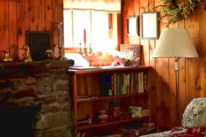 A cozy room with a stone fireplace, a bookshelf with various books, a lamp, and a window with curtains. - Enchanting Historic Cabin Haven near Black Hills Mountain in South Dakota (Keystone)
