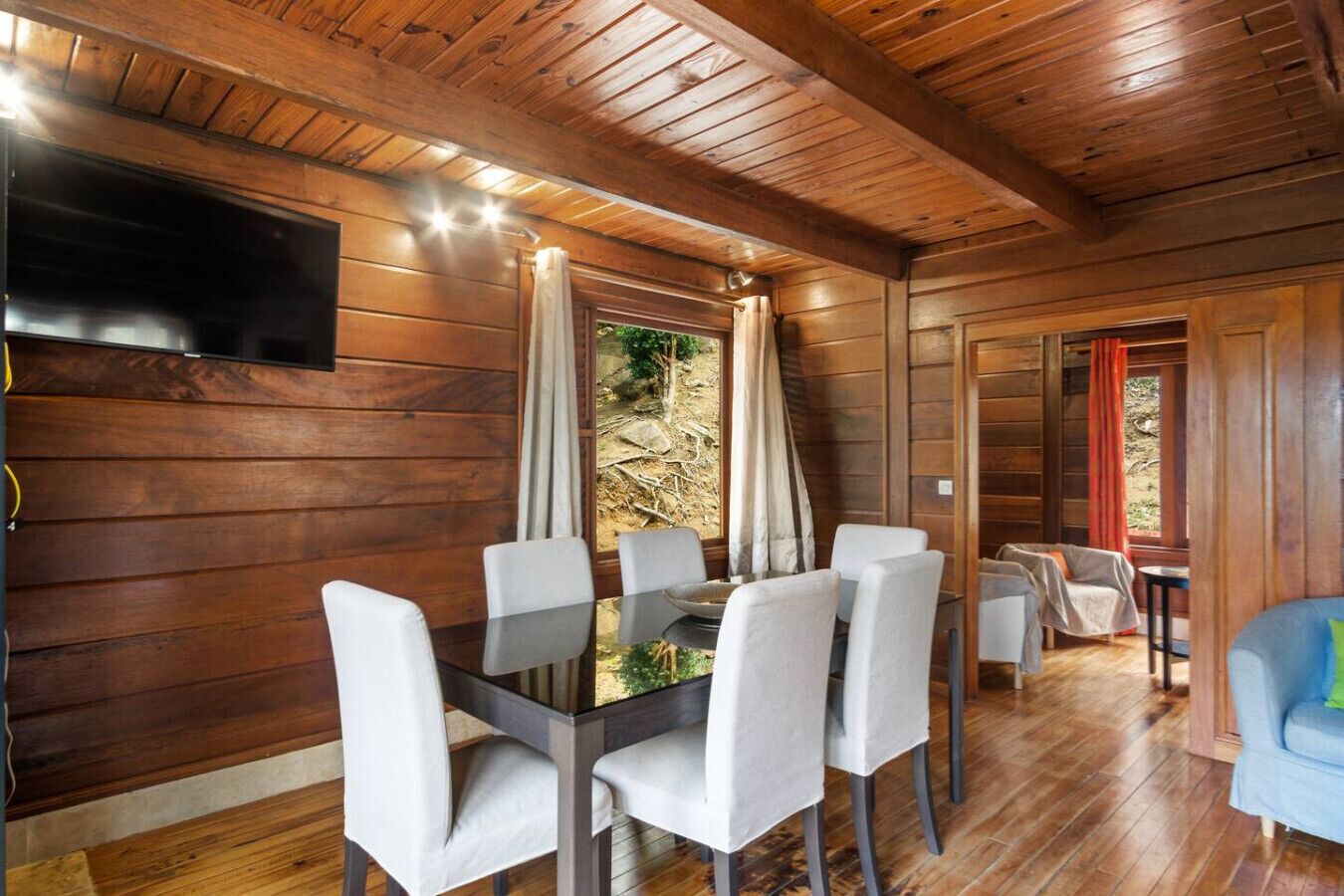A wooden interior with a dining area featuring a table and chairs, a television mounted on the wall, and a view of the outdoors through a window.