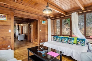 A wooden interior with a bed, a glass coffee table, and a view of the outdoors.