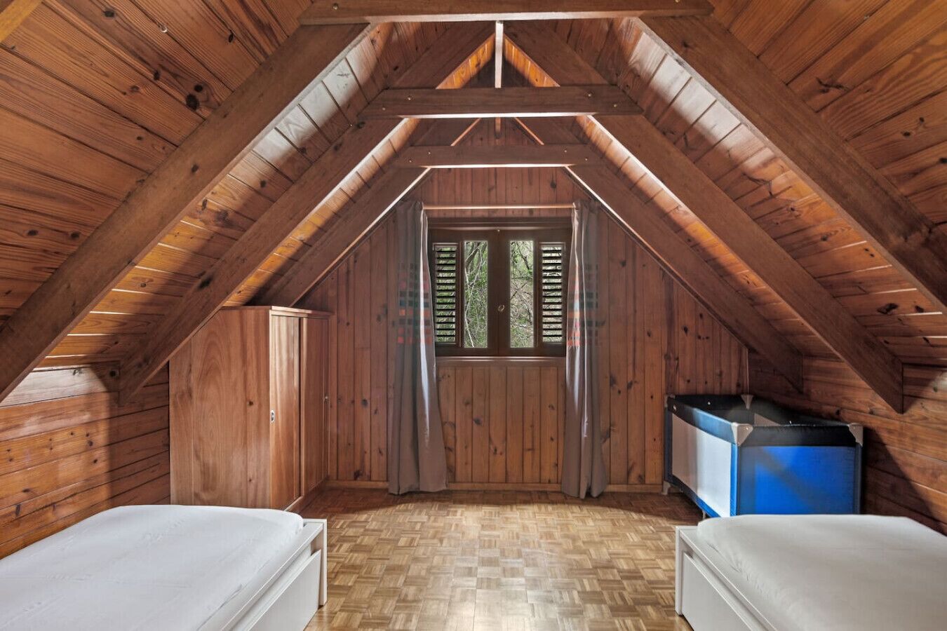 Um pequeno quarto de sótão com chão de madeira, teto inclinado, duas camas, uma janela com persianas e um armário azul.
