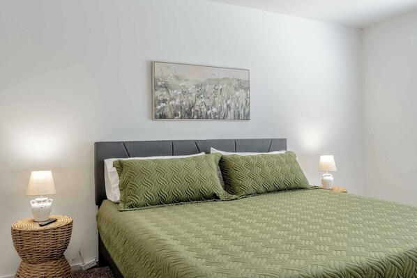 Una cama bien hecha con colcha verde, dos almohadas y un cabecero. Hay un cuadro en la pared sobre la cama, y dos lámparas de noche a cada lado.