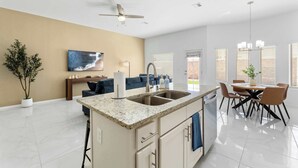 A modern kitchen with a central island, a dining area with a table and chairs, a ceiling fan, and a large window with blinds.