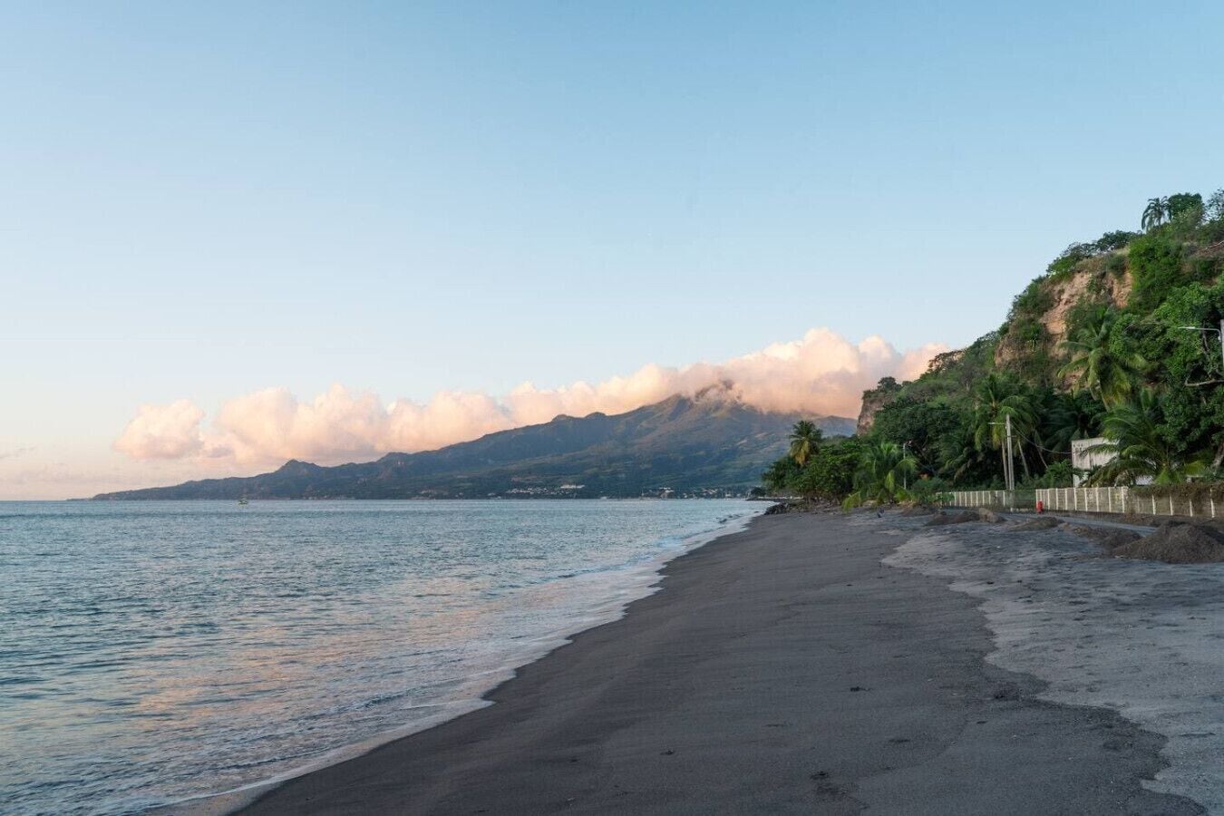 Uma praia com céu claro, mar calmo e montanha ao fundo.