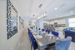 A modern dining area with a long table set for six, surrounded by blue chairs, and a kitchen in the background. - Bengal Court Reserve (Kissimmee)