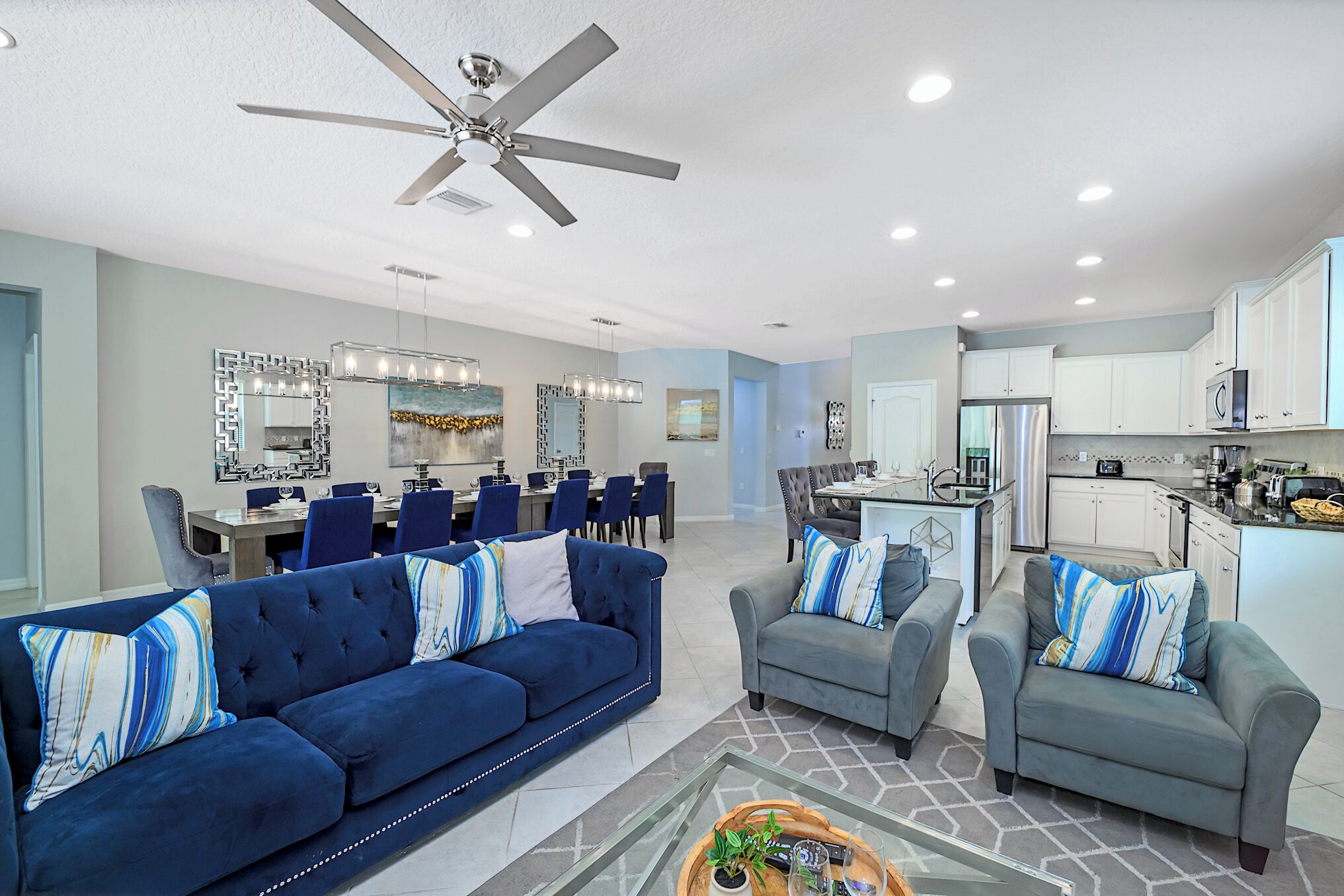 A modern living room with a blue sofa, grey armchairs, and a dining area with a chandelier.