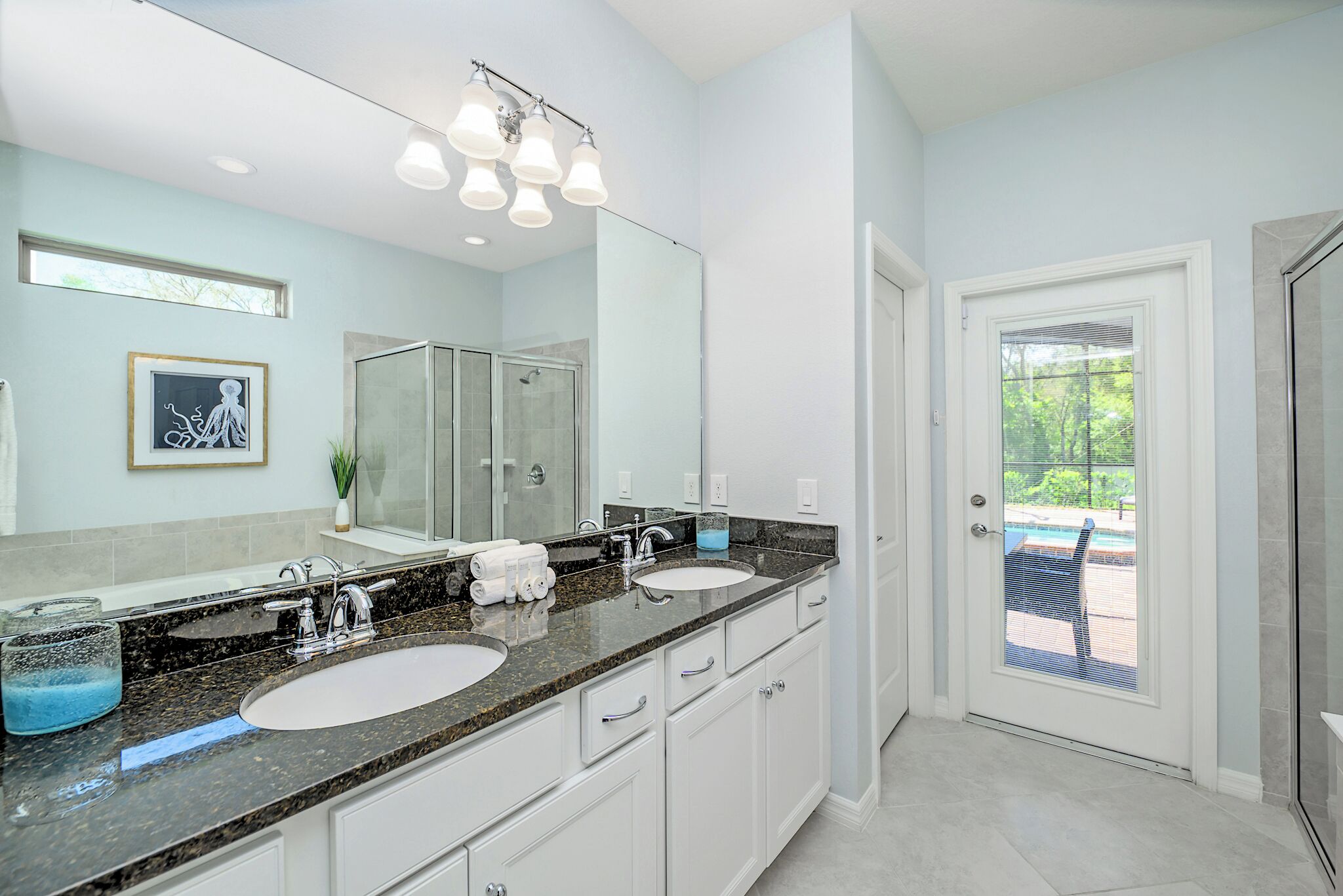 A modern bathroom with a large mirror, two sinks, a shower, and a door leading to a balcony.