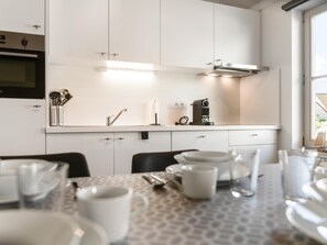 A modern kitchen with white cabinetry, a built-in oven, a coffee machine, and a dining area with a table set for two. - Cozy holiday home with a south-facing garden (Koksijde)