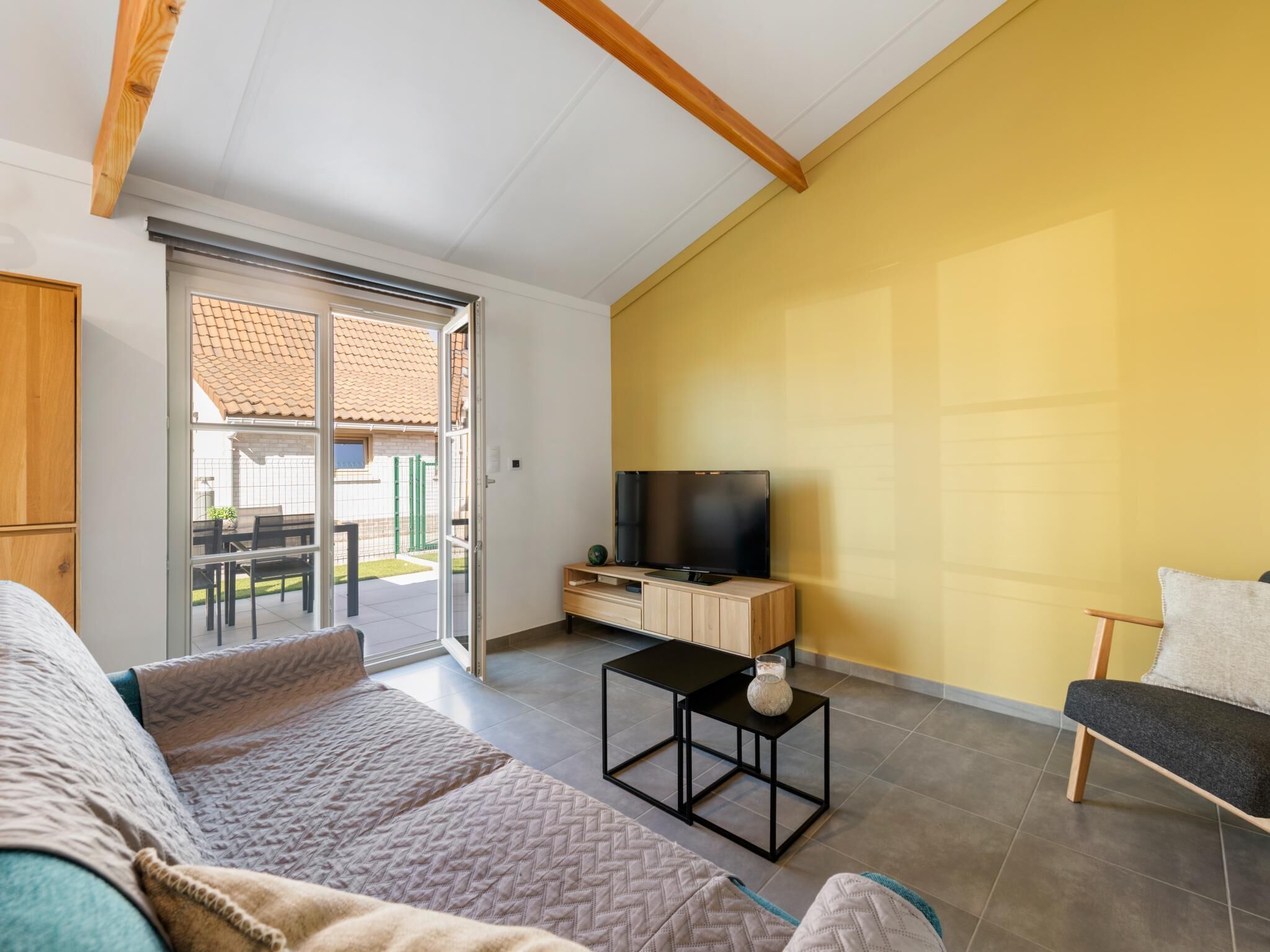 Een woonkamer met een bank, een televisie op een houten dressoir en een schuifpui naar een buitenruimte.