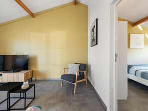 A modern living room with a flat-screen TV, a wooden coffee table, a grey armchair, and a bed with a patterned bedspread. - Cozy holiday home with a south-facing garden (Koksijde)