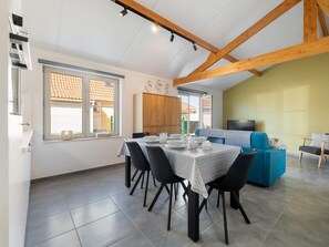 A dining area with a table set for four, a sofa, and a TV in a room with wooden beams and a tiled floor. - Cozy holiday home with a south-facing garden (Koksijde)