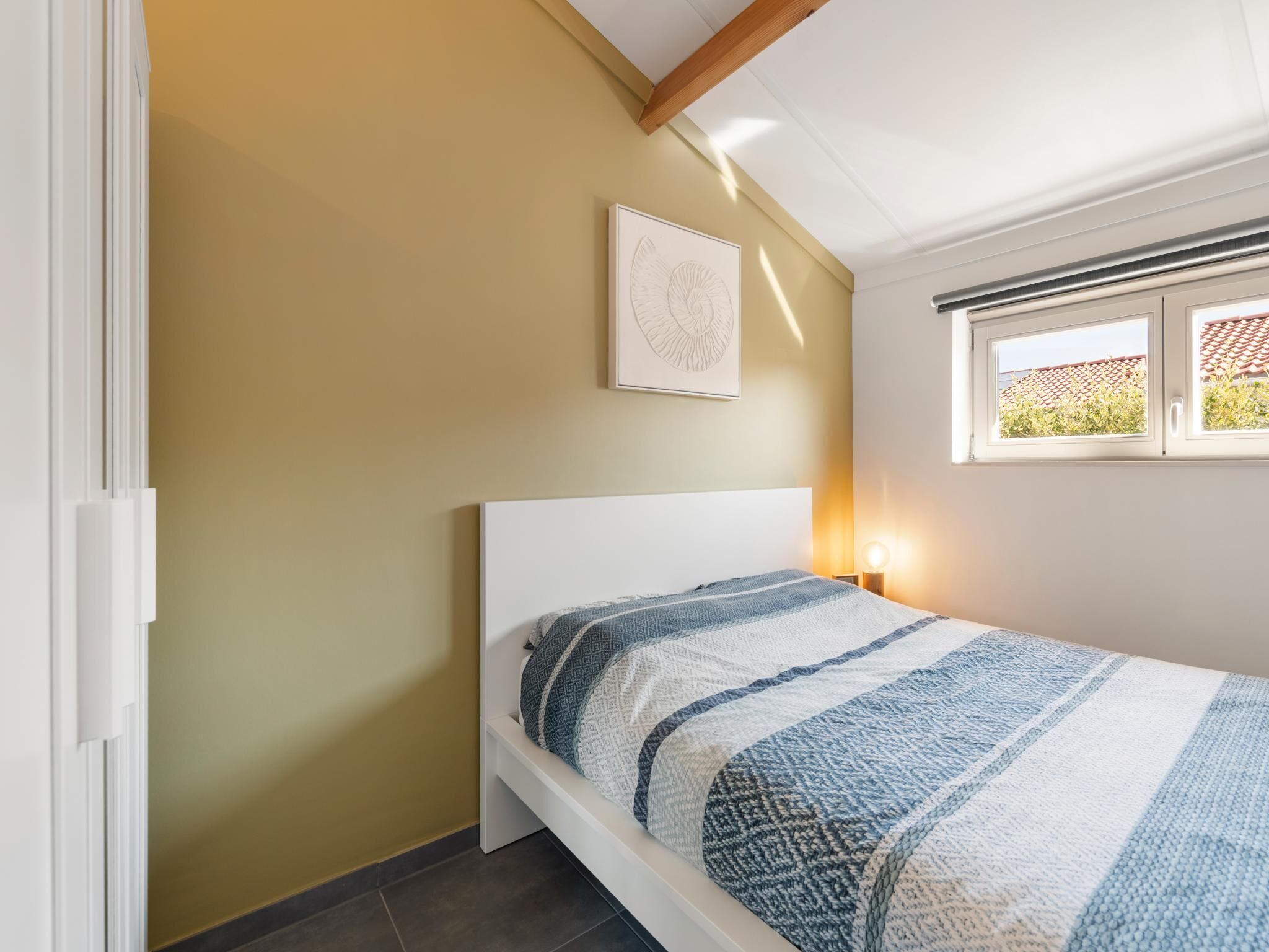 A bedroom with a single bed, a window with a view of greenery, and a framed picture on the wall.