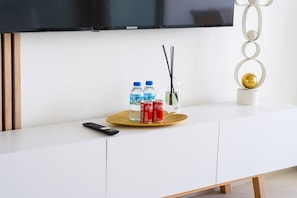 Ein modernes Wohnzimmer mit einem Flachbildfernseher, einer Fernbedienung, einer Ablage mit Wasserflaschen, einer Cola Dose und einem dekorativen Gegenstand.