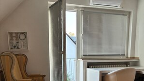 Et værelse med trægulv, et skrivebord med stol, en sofa og en skydedør, der fører til en balkon.