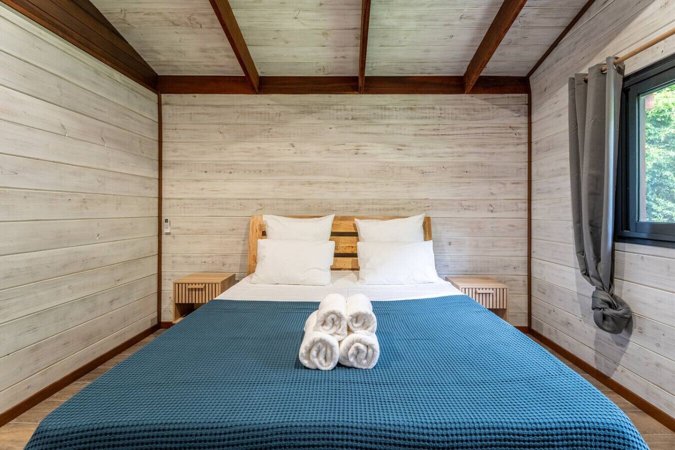Ein Zimmer mit einem Bett, Holzwänden, einem Fenster mit Vorhängen und einer blauen Bettdecke.