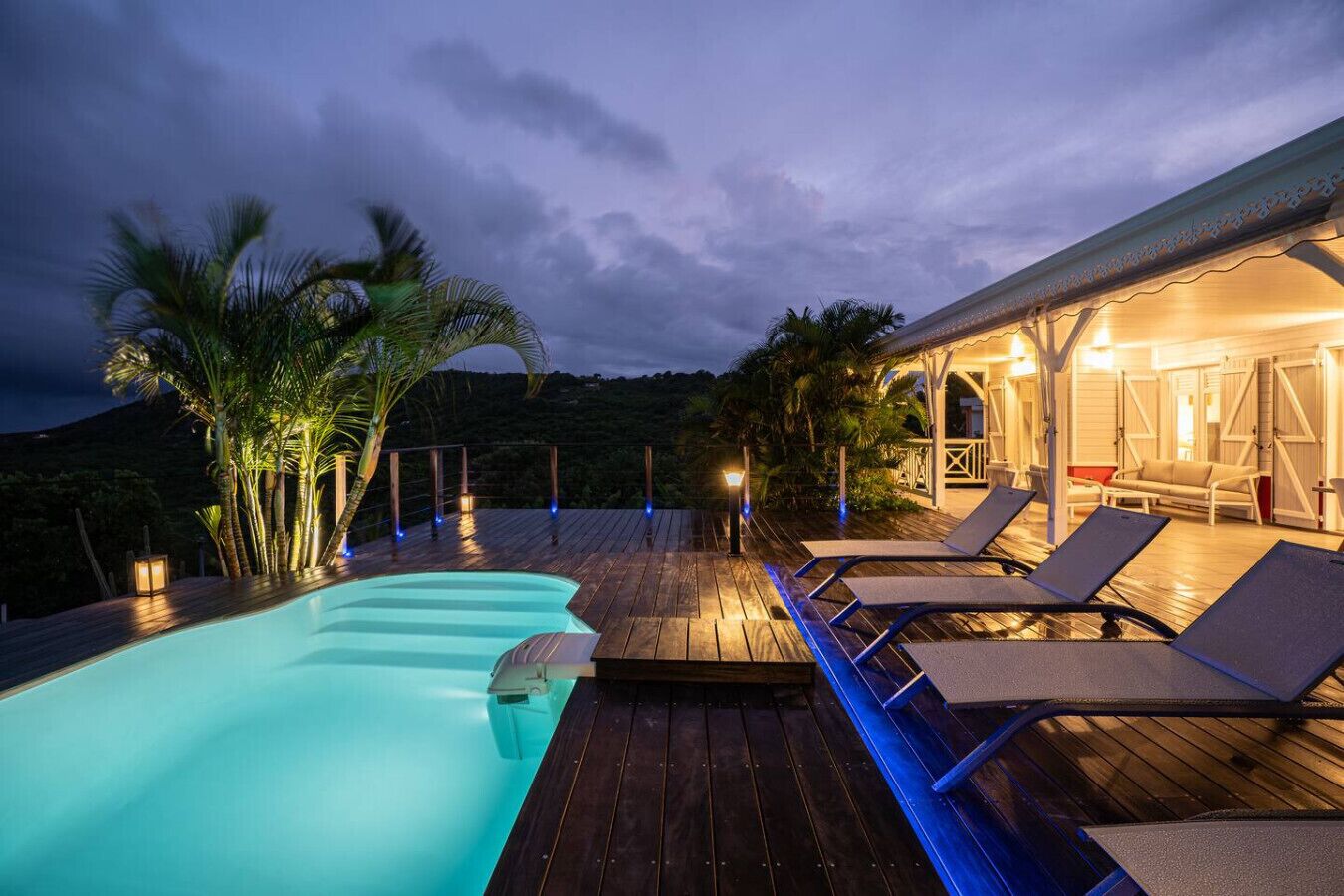 Un espace aménagé au bord de la piscine, avec des chaises longues et une terrasse en bois, entouré de palmiers et d’un bâtiment illuminé.
