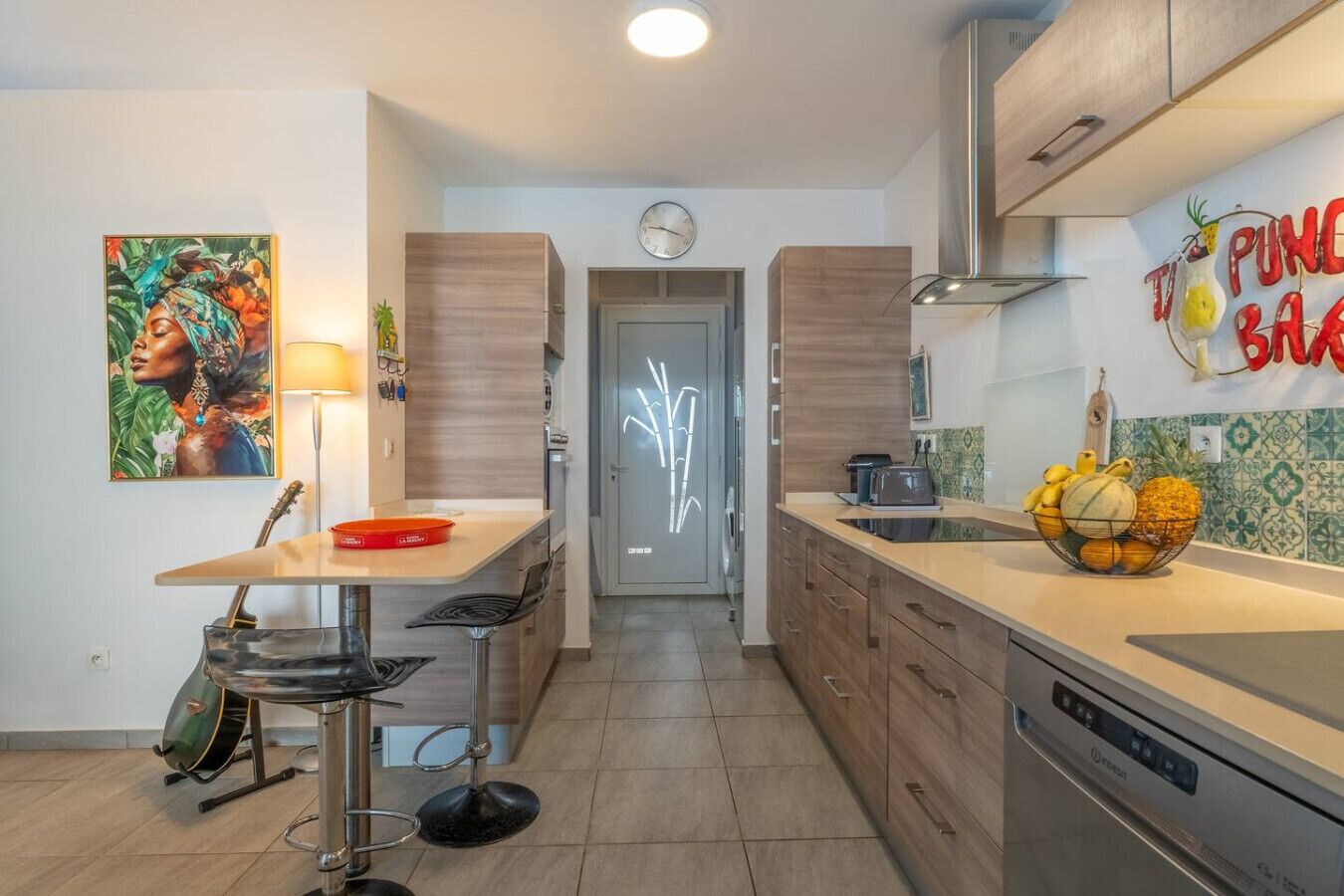 Une cuisine moderne avec un coin repas, une horloge et un panier de fruits sur le plan de travail.