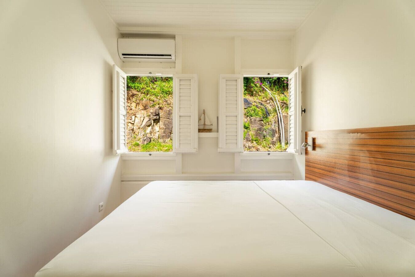 A bedroom with a bed, two windows with shutters, an air conditioning unit, and a wooden headboard.