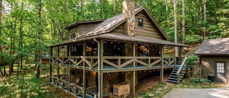 A rustic cabin with a wooden deck and a stone chimney, surrounded by dense forest.