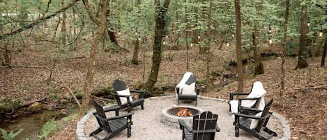 A circular seating arrangement with four Adirondack chairs around a central fire pit in a forested area.