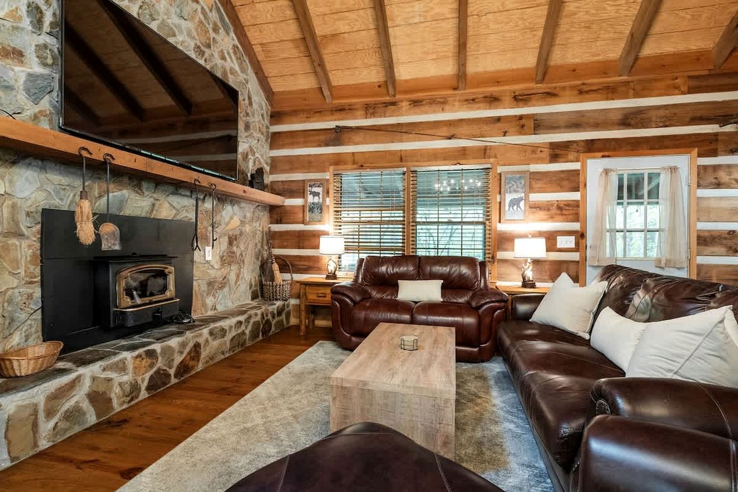 A log cabin interior with a stone fireplace, leather furniture, and wooden beams.