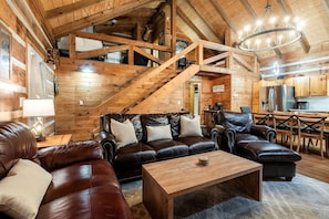 A spacious living area with a wooden staircase, leather sofas, a coffee table, and a kitchen in the background. - Enchanting Creekside Mountain Cabin Retreat with Luxury Amenities in the Heart of Scenic Blue Ridge, Georgia (Blue Ridge)