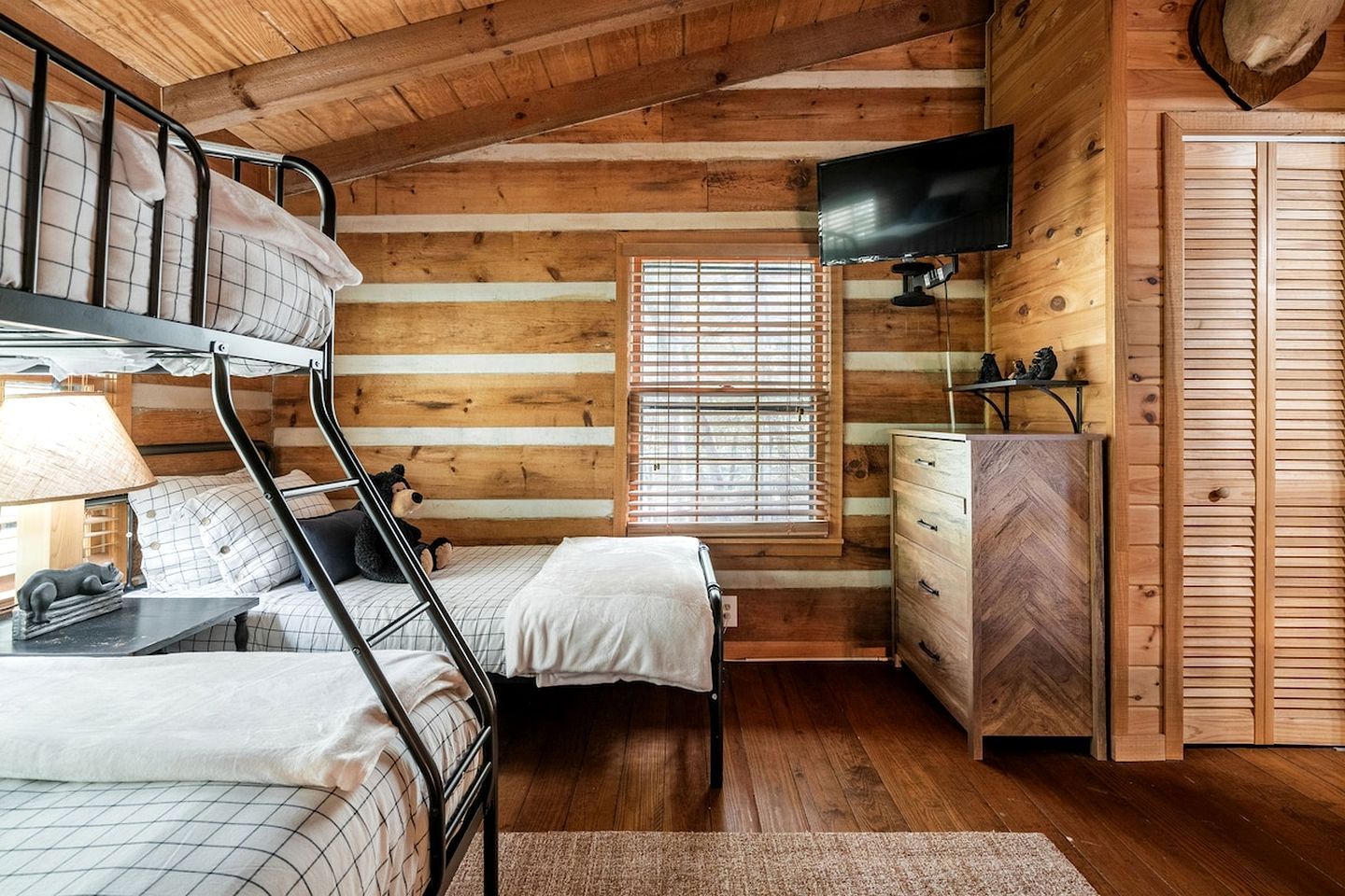 A cabin-style bedroom with a bunk bed, a dresser, a television, and a window with blinds.